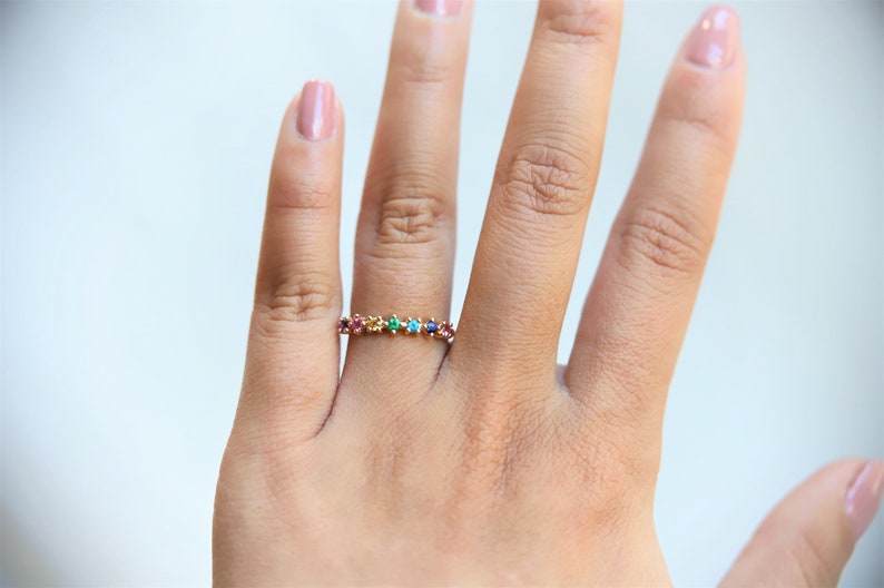 May include: A gold band ring with a row of colorful gemstones. The ring is on a person's finger.