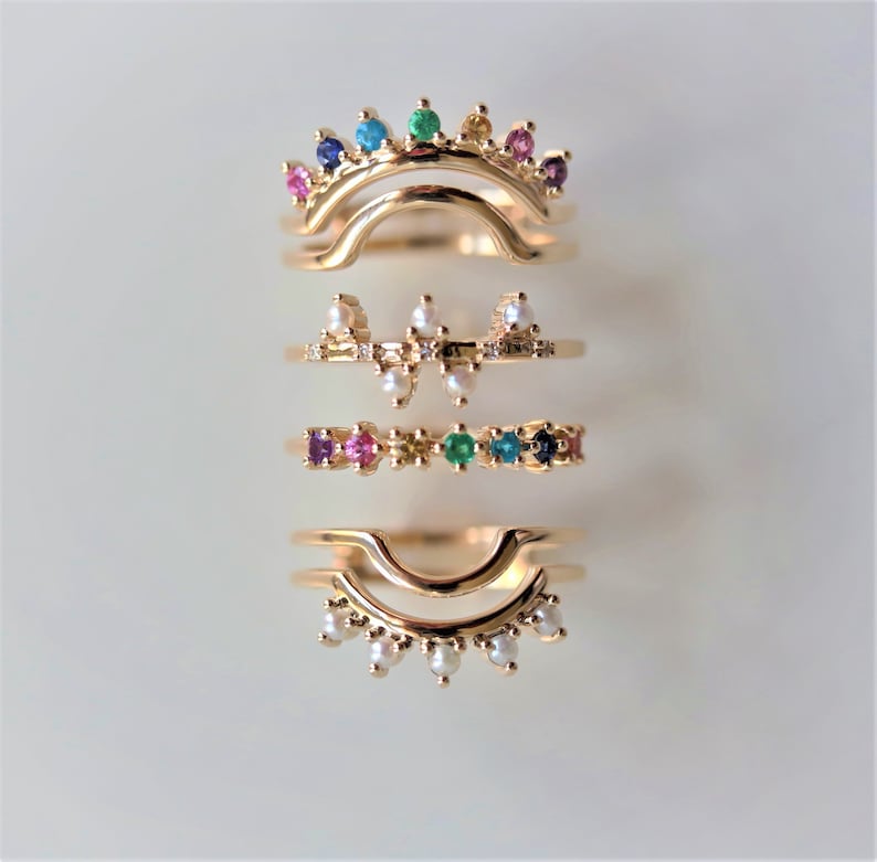 May include: Set of four gold rings with colorful gemstones and pearls. The rings are stacked on top of each other, with the top ring featuring a rainbow of gemstones.