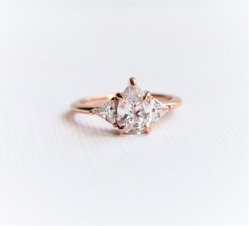 Puede incluir: Un anillo de compromiso de oro rosa con una piedra central grande, redonda y transparente y dos piedras triangulares m&aacute;s peque&ntilde;as y transparentes a cada lado.