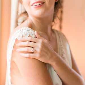 May include: A rose gold engagement ring with a starburst design featuring a central gemstone and surrounding smaller stones. The ring is on a hand, with a cream-colored lace-trimmed garment visible in the background. The image has a soft, warm color palette.