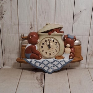 May include: A wall clock featuring three teddy bears, one peeking out of a ceramic pot, another holding a spoon, and the third sitting on a shelf. The clock face is white with black numbers and hands. The shelf is decorated with a blue and white checkered tablecloth.