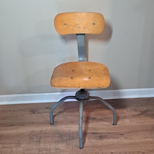 May include: A swivel chair featuring a light brown wooden seat and backrest attached to a gray metal frame with four legs. The chair has a central metal support column and visible rivets on the seat and backrest, showing signs of wear.