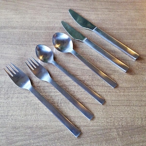 May include: A set of stainless steel flatware, including forks, spoons, and knives, arranged on a wooden surface. The utensils have a simple, modern design with rectangular handles.