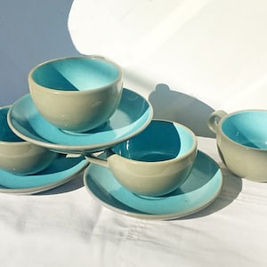 May include: Set of four ceramic teacups and saucers. The cups and saucers are a light gray color with a turquoise blue interior.