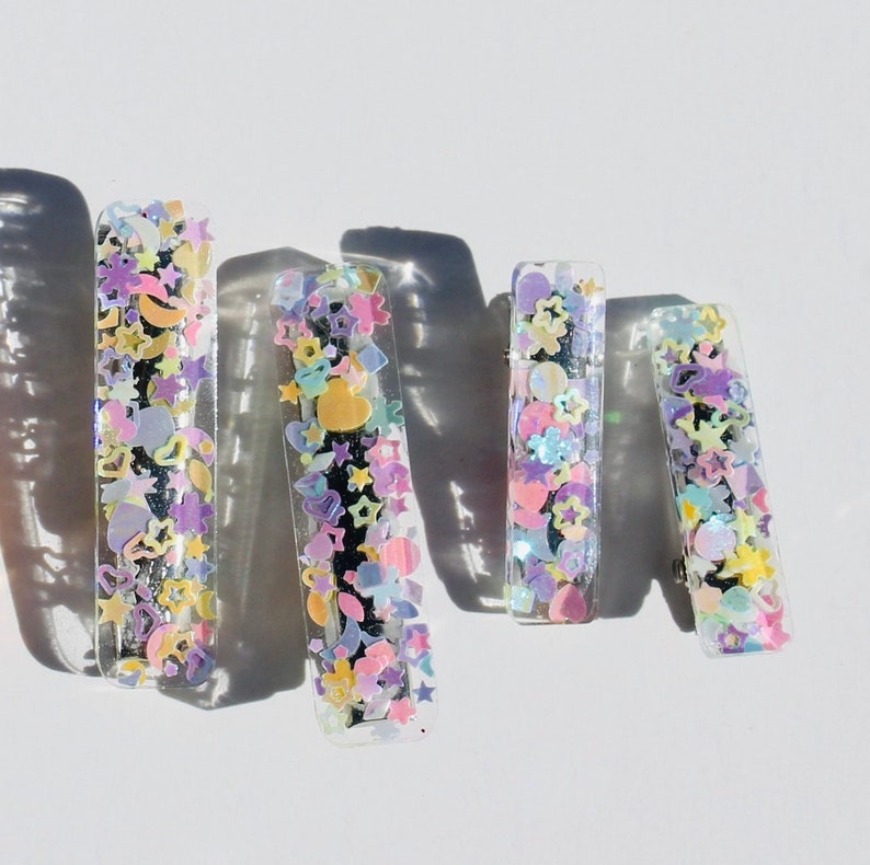 Op de afbeelding: Vier transparante haarclips met kleurrijk glitter en confetti erin. De clips zijn rechthoekig en hebben een glanzende afwerking.