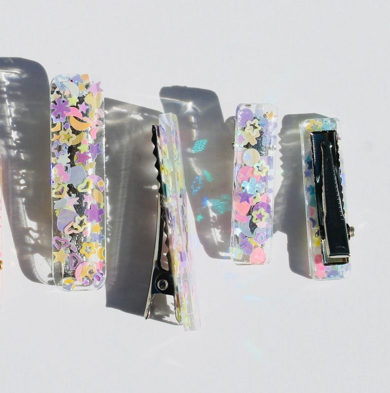 Op de afbeelding: Vier transparante haarclips met verschillende gekleurde glitters en iriserende pailletten erin. De clips zijn rechthoekig en hebben een zilveren metalen sluiting.