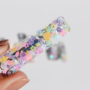 Op de afbeelding: Een transparante plastic haarclip met kleurrijk glitter en confetti erin. De glitter heeft de vorm van sterren, harten en andere kleine vormen.
