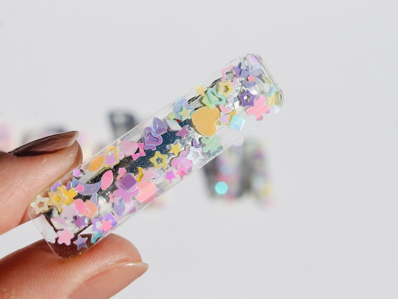 Op de afbeelding: Een transparante plastic haarclip met kleurrijk glitter en confetti erin. De glitter heeft de vorm van sterren, harten en andere kleine vormen.