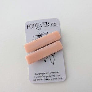 May include: Two rectangular, light peach-colored hair clips rest on a white card. The card features the text "FOREVER co." in black script, along with a floral illustration and additional text. The clips are smooth and have a subtle sheen.