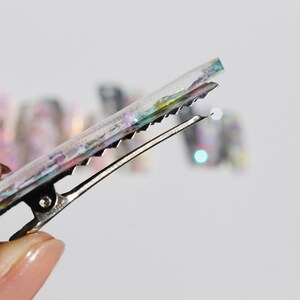 Op de afbeelding: Een transparante haarclip met iriserende glitter ingebed in de hars. De clip is zilver en heeft een textuur.