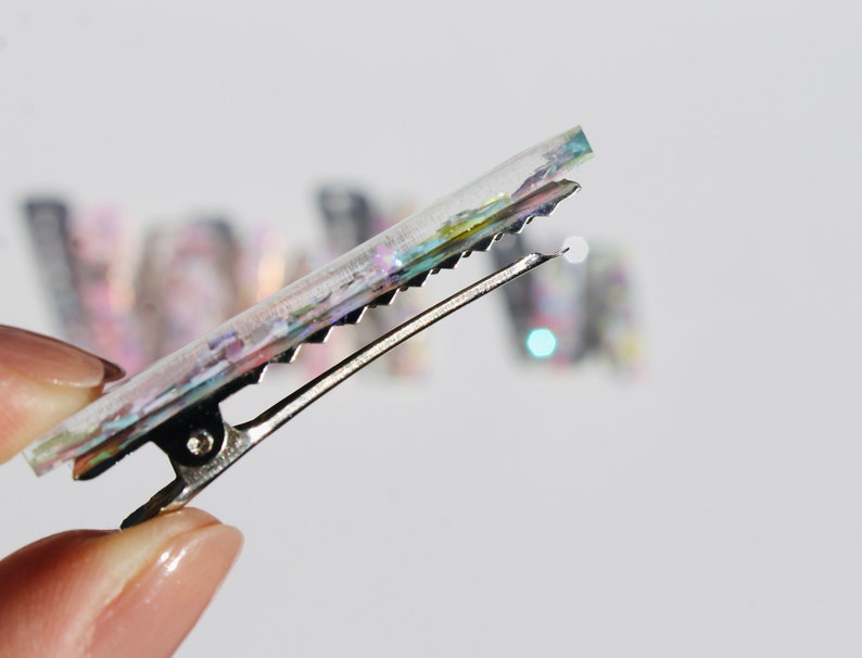 Op de afbeelding: Een transparante haarclip met iriserende glitter ingebed in de hars. De clip is zilver en heeft een textuur.