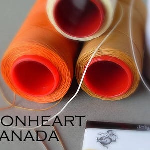 May include: Three spools of thread, one orange, one beige, and one white, with red plastic bobbins. The spools are labelled "Lionheart Canada".