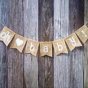 May include: A burlap banner with the words "oh baby!" in white lettering and a white heart. The banner is strung on twine and hangs against a rustic wooden background, perfect for a baby shower or nursery decor.