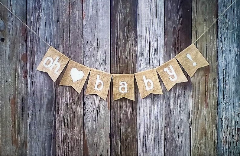 May include: A burlap banner with the words "oh baby!" in white lettering and a white heart. The banner is strung on twine and hangs against a rustic wooden background, perfect for a baby shower or nursery decor.