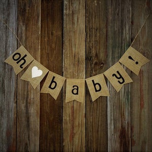 May include: A burlap banner with the words "oh baby!" in black letters and a white heart. The banner is strung on twine and hangs against a rustic wooden background, perfect for a baby shower.