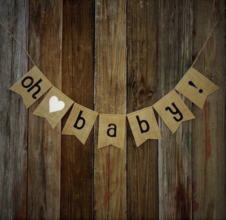 May include: A burlap banner with the words "oh baby!" in black letters and a white heart. The banner is strung on twine and hangs against a rustic wooden background, perfect for a baby shower.