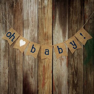 May include: A burlap banner with the words "oh baby!" in navy blue letters and a white heart. The banner is strung against a rustic wooden background, perfect for a baby shower or nursery decor.