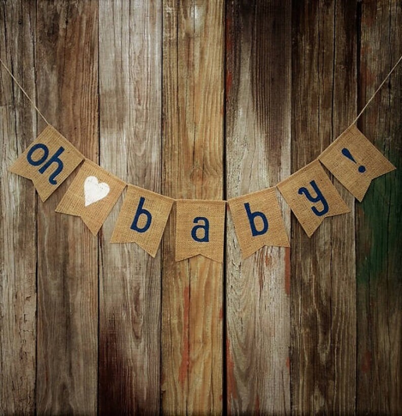 May include: A burlap banner with the words "oh baby!" in navy blue letters and a white heart. The banner is strung against a rustic wooden background, perfect for a baby shower or nursery decor.