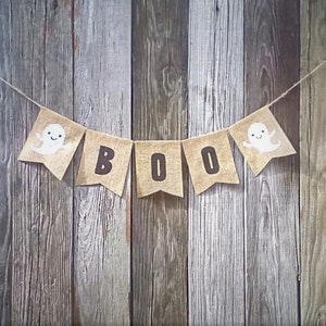 May include: A burlap banner with the word "BOO" in black letters and ghost illustrations on each end, hanging against a weathered wood background.