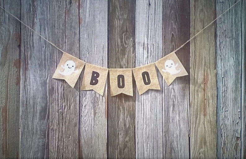 May include: A burlap banner with the word "BOO" in black letters and ghost illustrations on each end, hanging against a weathered wood background.