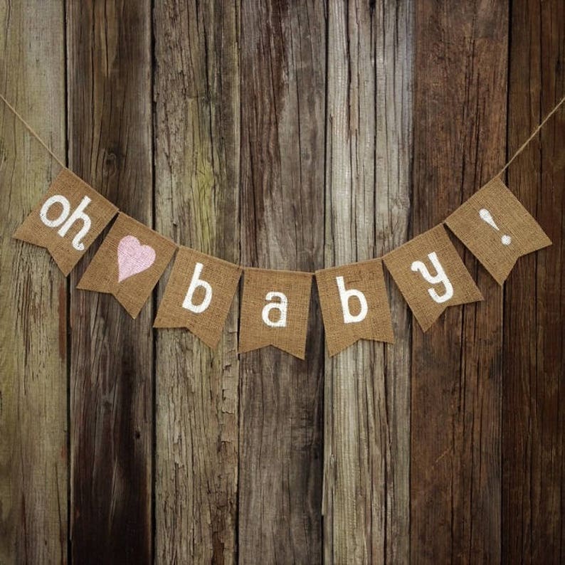 May include: A burlap banner with the words "oh baby!" in white lettering and a pink heart. The banner is strung on twine and hangs against a rustic wooden background. Perfect for a baby shower or nursery decor.
