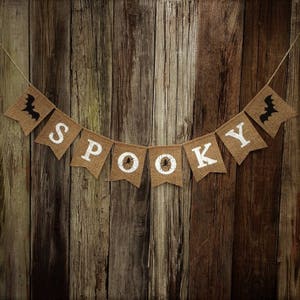 May include: A burlap banner with the word "SPOOKY" in white letters. The banner is decorated with black bats and spiders. The banner is hanging on a rustic wooden background.