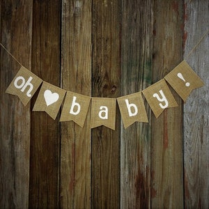 May include: A burlap banner with the words "oh baby!" in white letters. The banner includes a white heart symbol. The banner is strung on a thin twine and hangs against a wooden background.