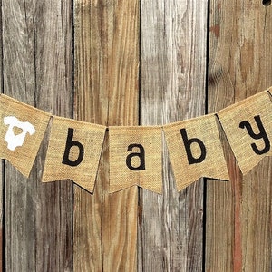May include: A burlap banner with the words "oh baby!" in black letters. The banner includes a white onesie graphic. The banner is strung on a thin wire and hangs against a rustic wooden background.