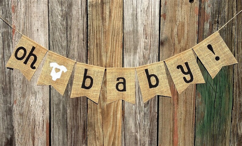 May include: A burlap banner with the words "oh baby!" in black letters. The banner includes a white onesie graphic. The banner is strung on a thin wire and hangs against a rustic wooden background.