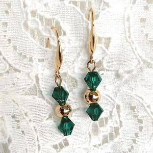 May include: A pair of gold-colored drop earrings. Each earring features a gold-colored hook, a faceted emerald green crystal, a gold-colored bead, and another emerald green crystal. The earrings are displayed on a white lace surface.
