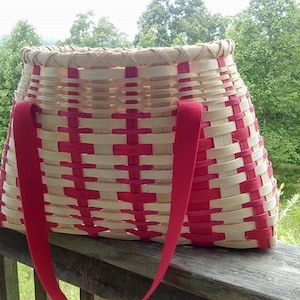 Puede incluir: Una cesta tejida con rayas rojas y blancas y asas rojas. La cesta está sentada en una barandilla de madera.