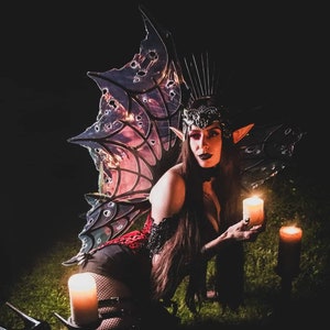 May include: A woman with long black hair and pointed ears wears a black and red corset, black fishnet stockings, and black boots. She holds a lit candle in her hand and is kneeling in front of two lit candles on the ground. She is wearing a large, iridescent butterfly wing costume with a black crown.