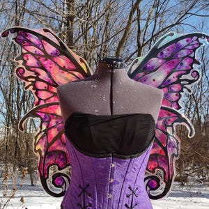 May include: A purple corset with black detailing and lacing, displayed on a mannequin. The corset is paired with a black strapless top. Large, colourful butterfly wings in shades of pink, purple, and black are attached to the mannequin's back. The background shows a snowy outdoor scene.