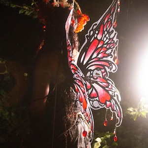 May include: Decorative butterfly wings in red and black, with dangling red jewels. The wings are attached to a dark garment. A gold crown and floral accents are visible at the top. The image is set against a dark background.