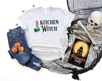 May include: A gray t-shirt with the text "Kitchen Witch" and a green potion bottle graphic. The shirt is displayed with a pair of blue jeans, a Halloween party invitation, and a skull and crossbones.