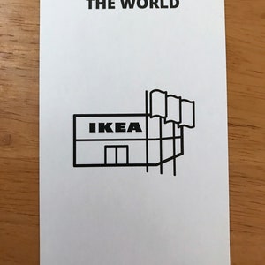 Puede incluir: Una tarjeta rectangular blanca con esquinas redondeadas que presenta el texto negro THE WORLD en la parte superior y el n&uacute;mero romano xxi en la parte inferior. En el centro hay un dibujo lineal negro de una tienda IKEA con tres banderas.