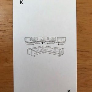 Puede incluir: Una carta de juego rectangular blanca con la letra K en las esquinas superior izquierda e inferior derecha. El centro muestra un dibujo lineal negro de un sof&aacute; seccional con seis cojines capitonados flotando encima.