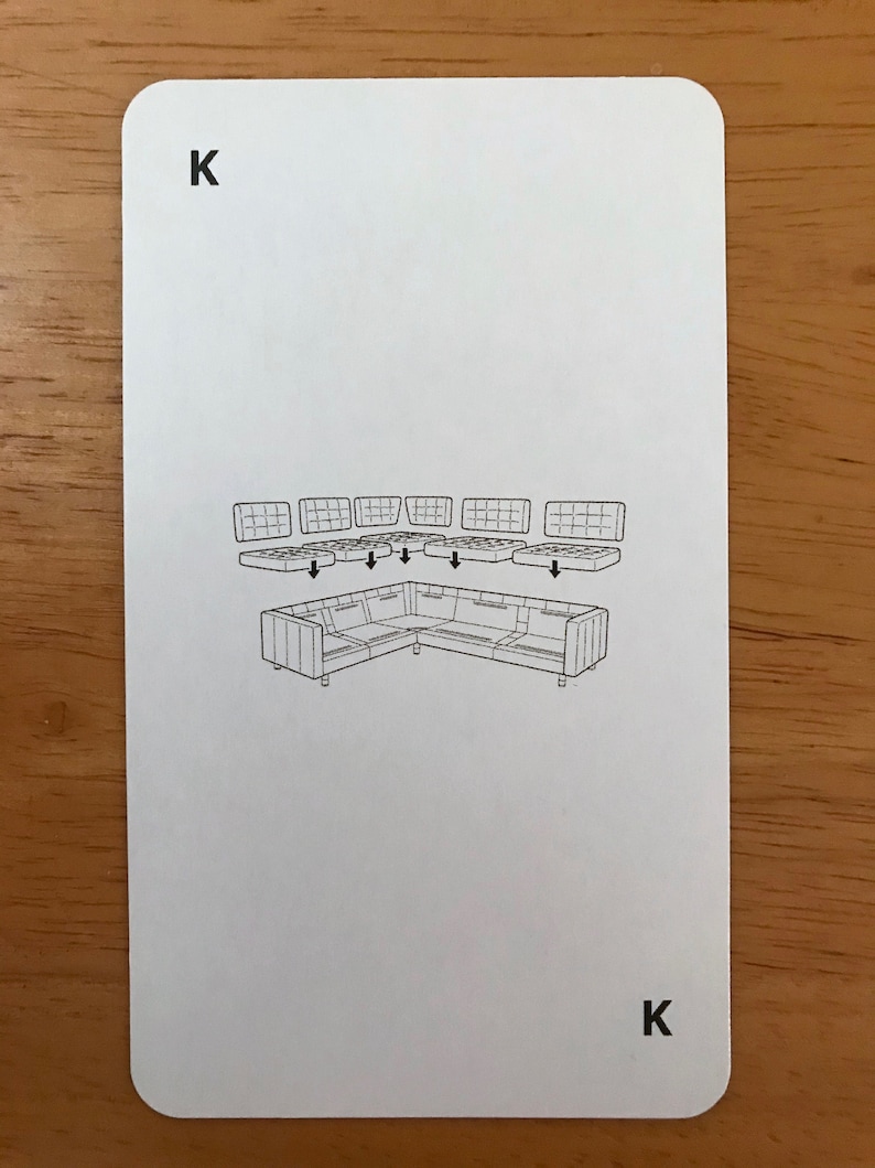 Puede incluir: Una carta de juego rectangular blanca con la letra K en las esquinas superior izquierda e inferior derecha. El centro muestra un dibujo lineal negro de un sof&aacute; seccional con seis cojines capitonados flotando encima.