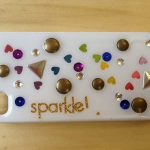 Puede incluir: Una funda de teléfono rectangular blanca decorada con varios adornos, incluyendo tachuelas doradas, acentos triangulares plateados, lentejuelas coloridas y pequeñas formas de corazón. La palabra sparkle! está escrita en letras de purpurina dorada.