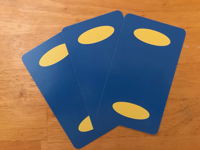Puede incluir: Tres cartas de juego rectangulares con esquinas redondeadas, con un fondo azul s&oacute;lido y dos formas ovaladas amarillas posicionadas en la parte superior e inferior de cada carta, dispuestas en un patr&oacute;n de abanico.