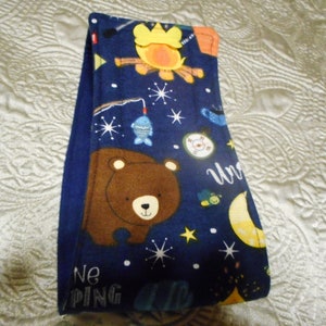 May include: A blue fabric headband with a camping theme. The headband features a brown bear, a campfire, a fish, a compass, a moon, and stars. The text "We're Camping" is printed on the headband.