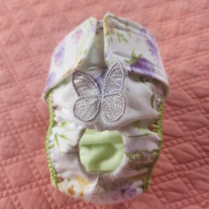 May include: A white cloth diaper with a floral pattern featuring purple and yellow flowers, green stems, and a silver embroidered butterfly appliqué on the front. The diaper has lime green trim along the leg openings and hook-and-loop fasteners.