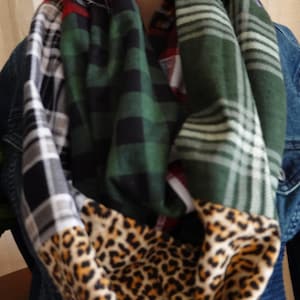 May include: A patchwork infinity scarf featuring a variety of plaid patterns in red, green, black, and white, with a leopard print accent. The scarf is made of soft fabric and is designed to be worn around the neck.