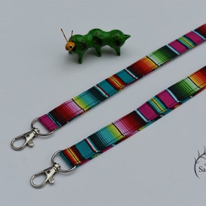 May include: Two colorful striped lanyards with silver swivel clips. The lanyards are made of fabric with a pattern reminiscent of a Mexican serape. The lanyards are on a white background.