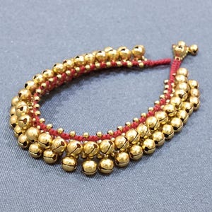 May include: Gold-colored bell anklet strung on a red cord. The anklet features multiple rows of small, round bells, creating a layered effect. The bells have a shiny finish, and the red cord provides a contrasting color.