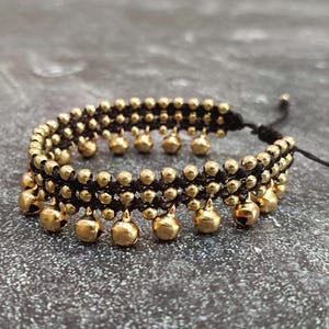 May include: A black and gold beaded bracelet with dangling gold-colored bells. The bracelet is made of black cord and small gold beads, with larger bell-shaped charms hanging from the bottom.