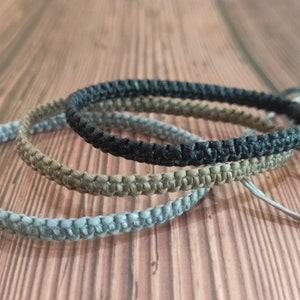 May include: Three braided bracelets in grey, brown, and black. The bracelets are made of a woven cord material.