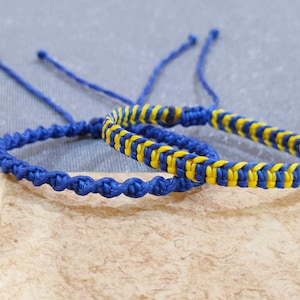 May include: Two adjustable macrame bracelets. One is solid blue, the other features a woven pattern of blue and yellow. The bracelets are displayed on a textured, neutral-toned surface, with a grey background. The bracelets are made of a woven cord.