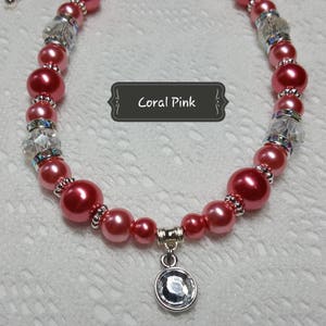 K&ouml;nnte beinhalten: Eine korallenrosa Perlenkette mit klaren Kristallperlen und einem silbernen Anh&auml;nger mit einem klaren Kristall. Die Kette tr&auml;gt den Text "Coral Pink" auf einem schwarzen Etikett.