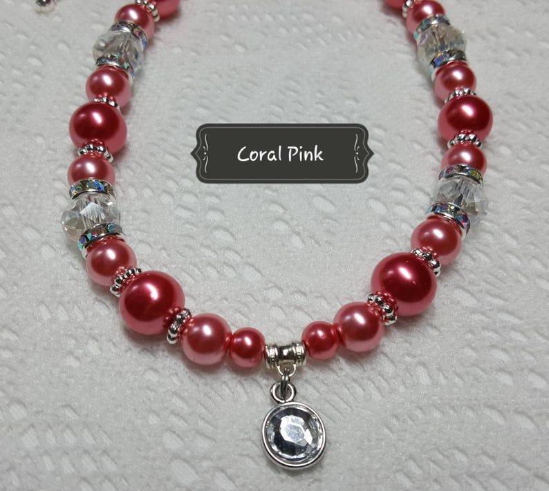 K&ouml;nnte beinhalten: Eine korallenrosa Perlenkette mit klaren Kristallperlen und einem silbernen Anh&auml;nger mit einem klaren Kristall. Die Kette tr&auml;gt den Text "Coral Pink" auf einem schwarzen Etikett.